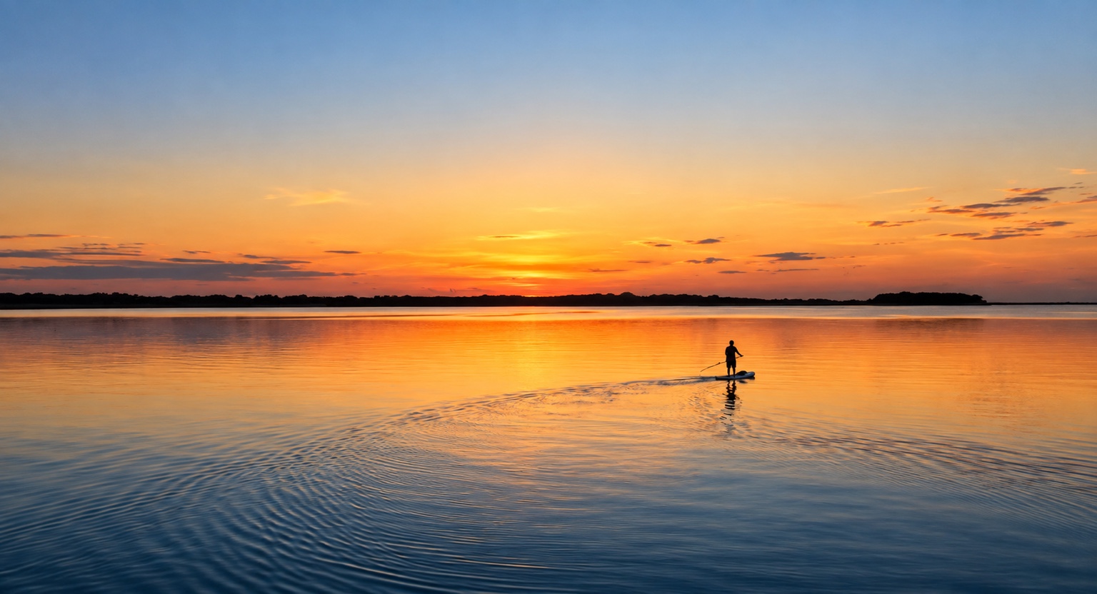 A paddleboarder crossing still water beneath a glowing orange sunset