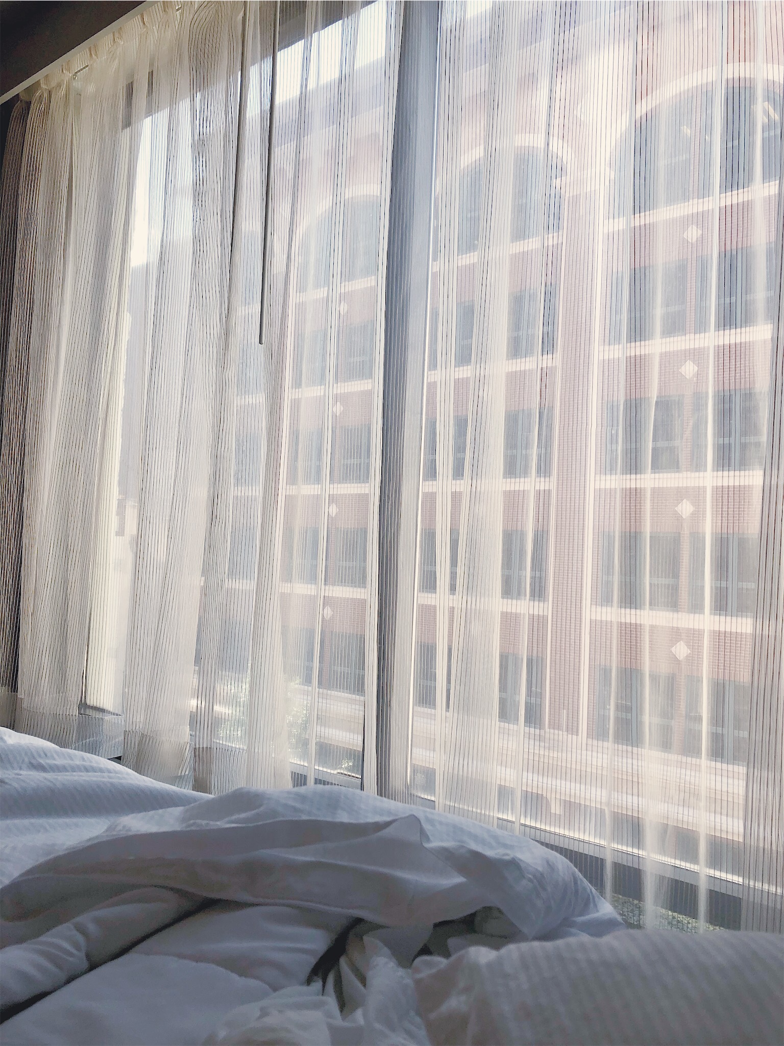 Unmade white hotel bedding beside sunlit sheer curtains